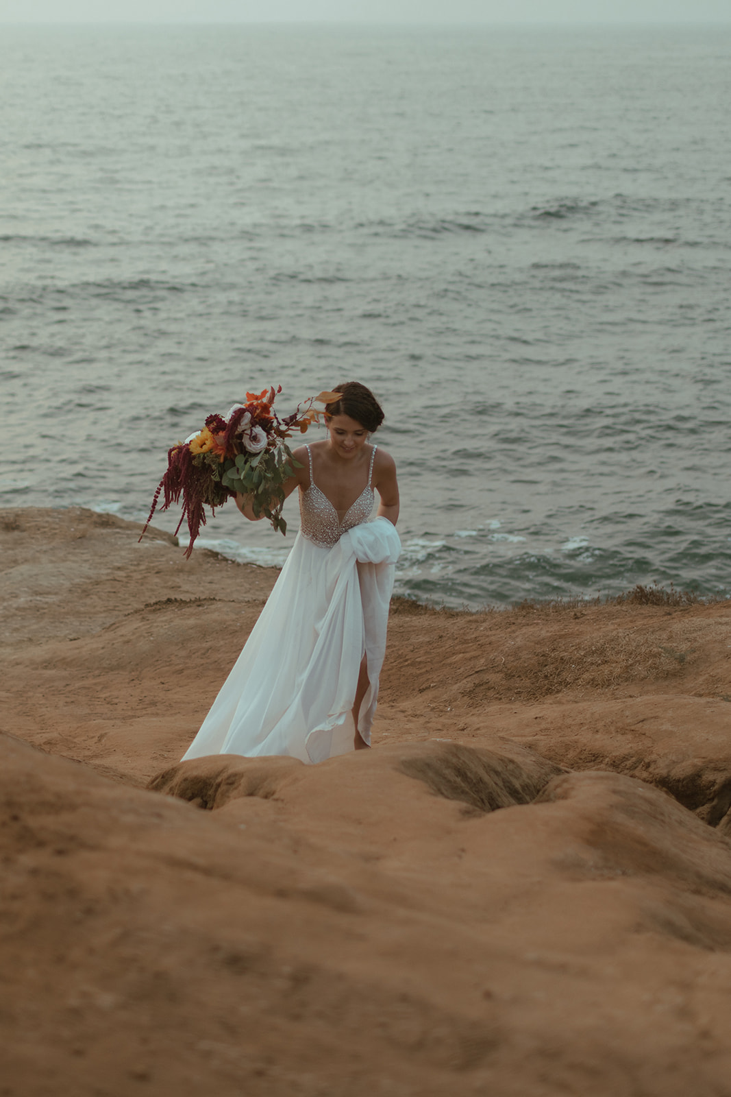 bride walking on san diego beach cliffside holding her boho wedding flowers