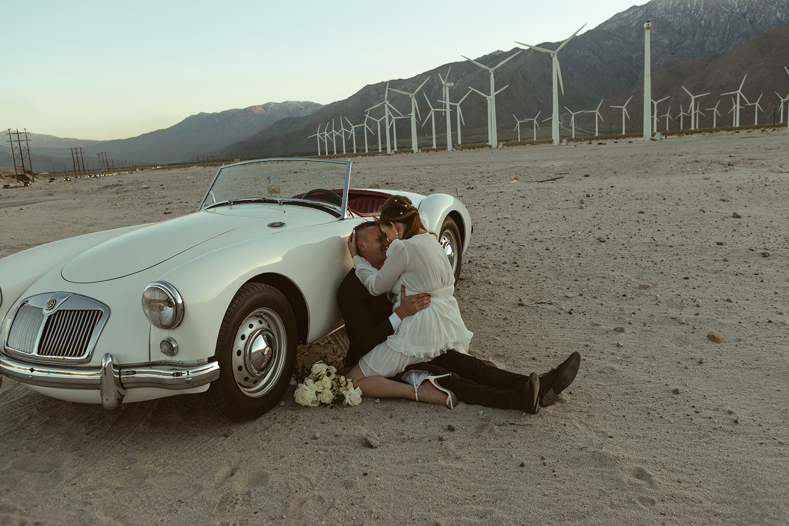 Retro Palm Springs Elopement with couple sitting on ground hugging in front of windmills